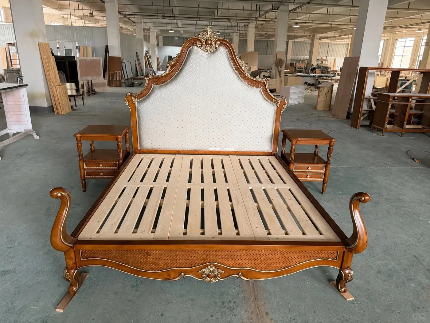 Factory floor photo of an ornate carved solid wood bed frame with upholstered headboard
