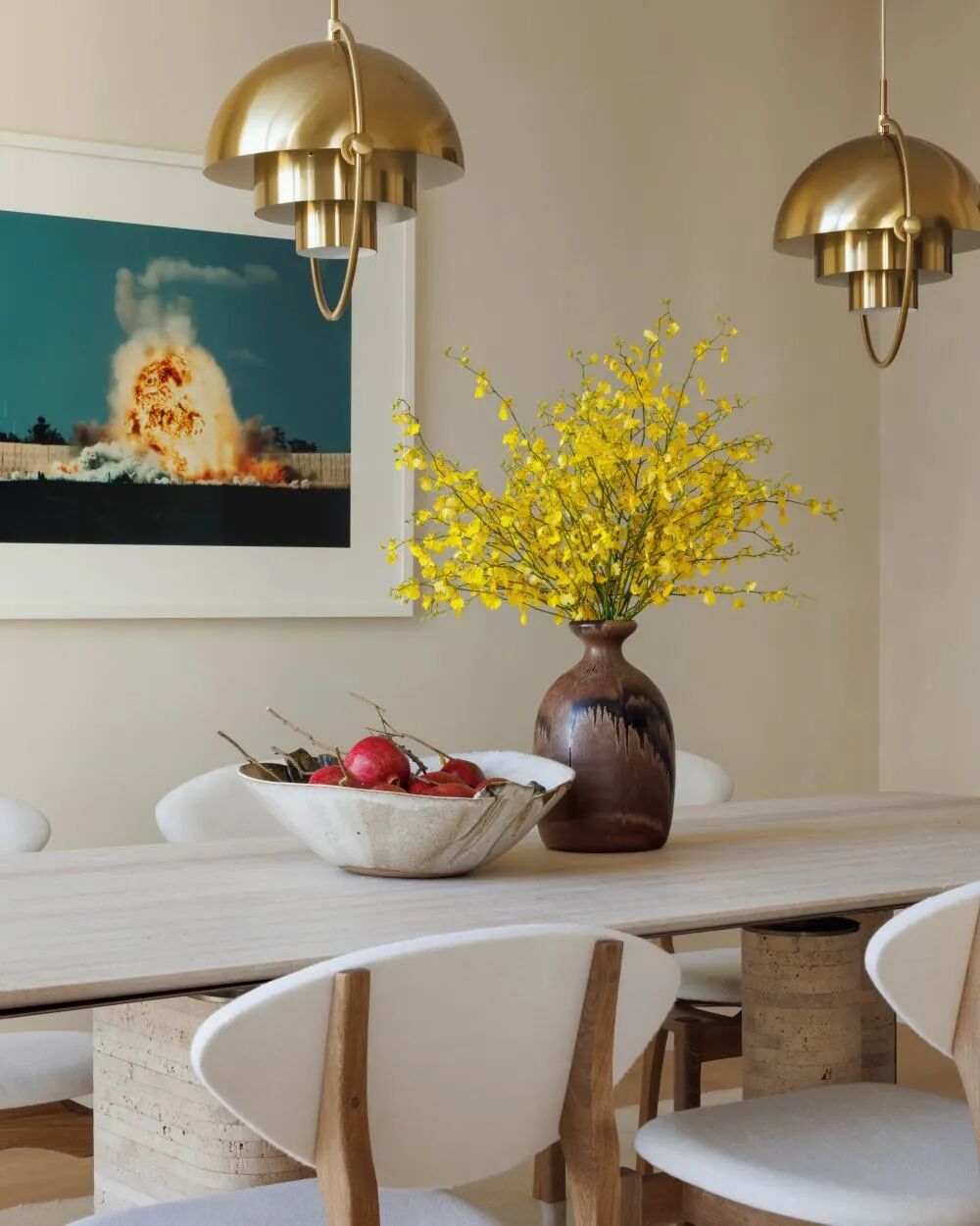 Close-up of a modern dining table with brass pendant lighting, yellow flowers, white chairs, and framed wall art