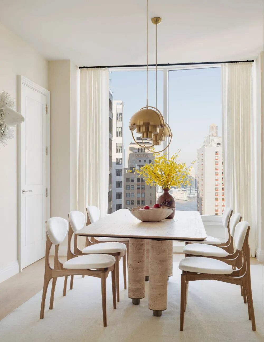 Bright modern dining room with sculptural wood table, white upholstered chairs, brass pendant lights, and city view