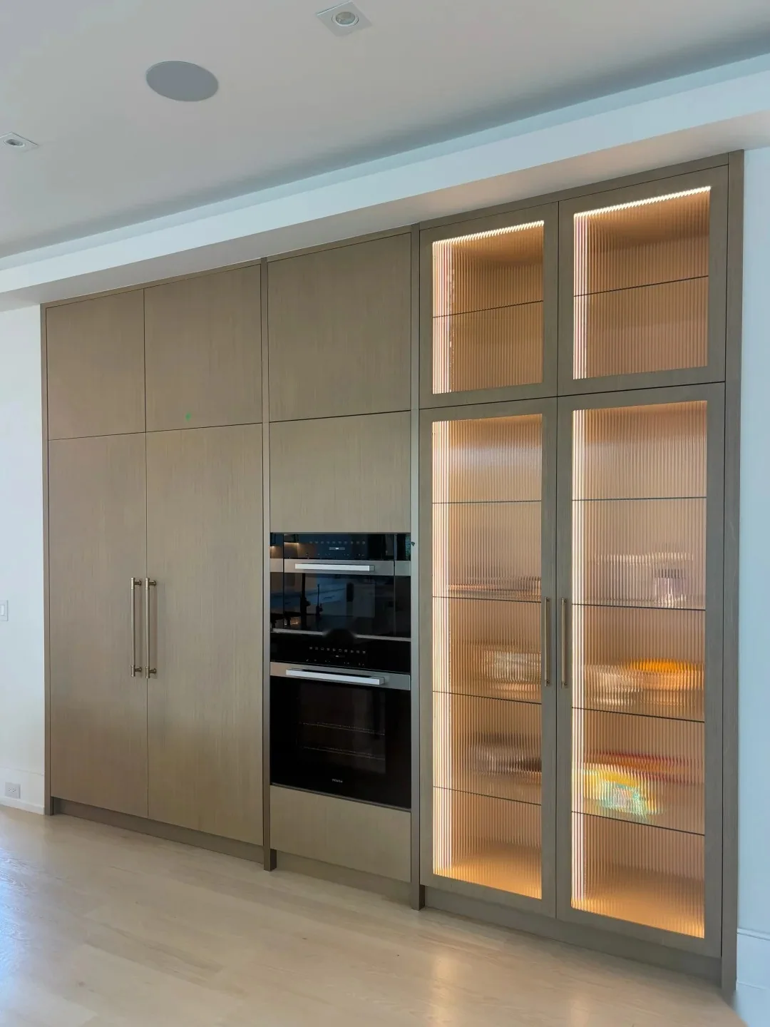 Full-height custom cabinetry with integrated ovens, tall storage, fluted glass doors and interior lighting