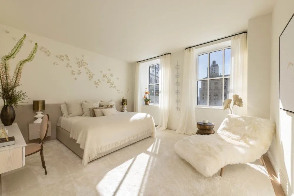 Sunlit neutral bedroom with soft white bedding, sheer curtains, city windows, and a cozy lounge chair