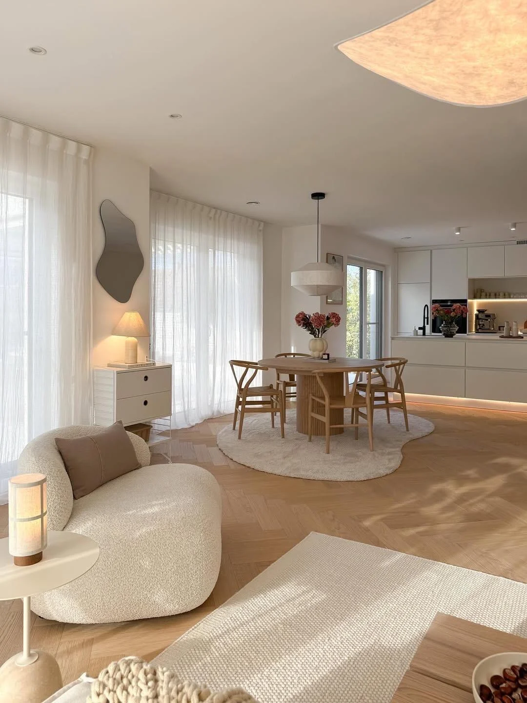 Open-plan Scandinavian dining space with rounded wood table, simple chairs and daylight filtering through sheer curtains